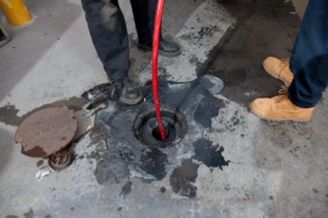 Two people stand near an open manhole; one inserts a red hose into the hole, with a metal cover and dark stains visible on the concrete ground.