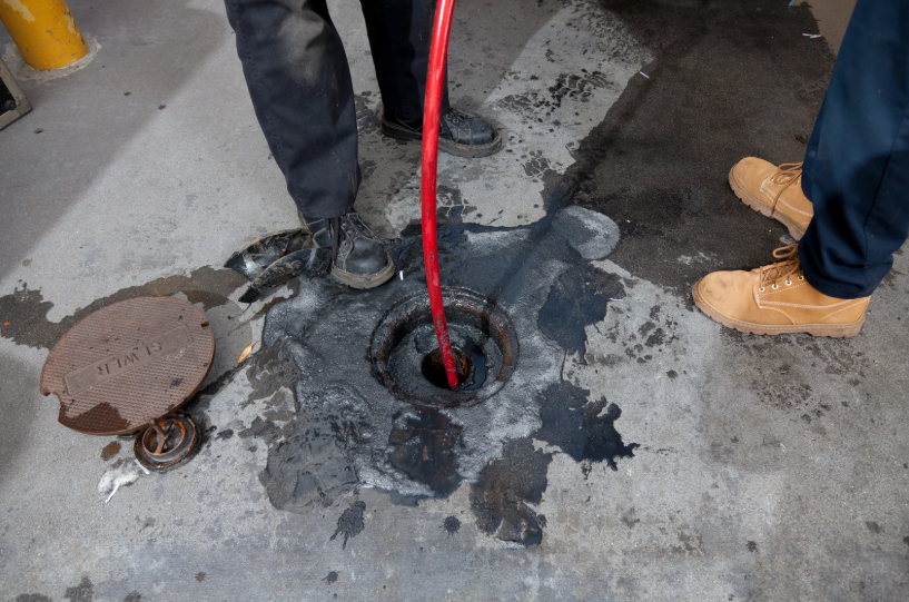 Two people stand near an open manhole; one inserts a red hose into the hole, with a metal cover and dark stains visible on the concrete ground.