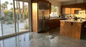A modern kitchen with wooden cabinets and a stainless steel refrigerator. A rag and a puddle of water are on the polished concrete floor near the refrigerator.