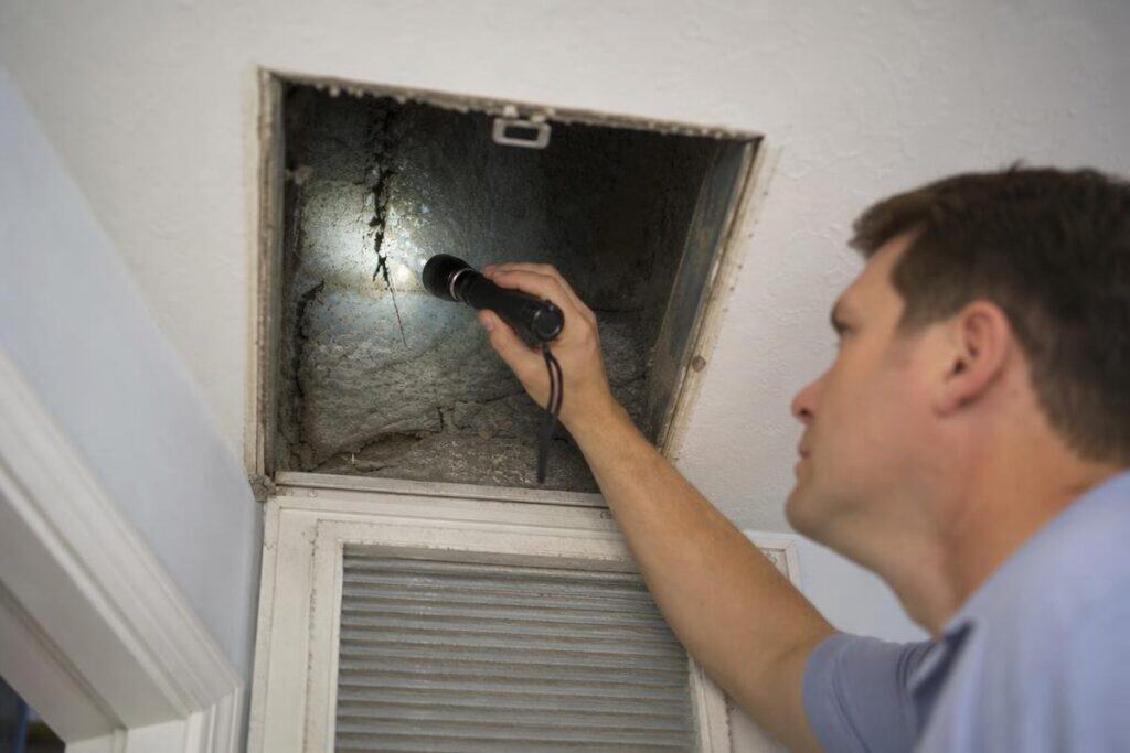 Man using a flashlight to inspect an open ceiling vent with visible dust and debris inside.