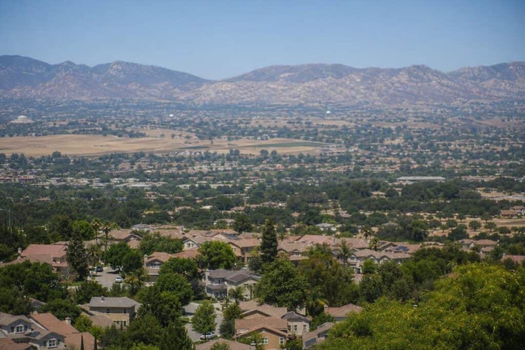 Suburban neighborhood with houses and trees in the foreground, wide farmland in the distance, and mountains under a clear blue sky.