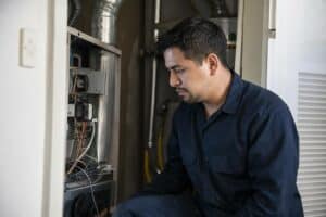 A man in a navy work uniform inspects the open panel of a furnace, examining the internal wires and components in a utility room.