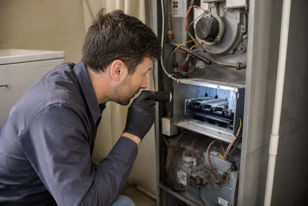 A man using a flashlight inspects the inside of an open furnace, wearing black gloves, with exposed wires and components visible.