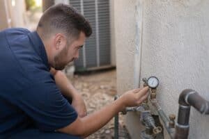 A man in a blue shirt checks a pressure gauge attached to a pipe on the exterior wall of a building.