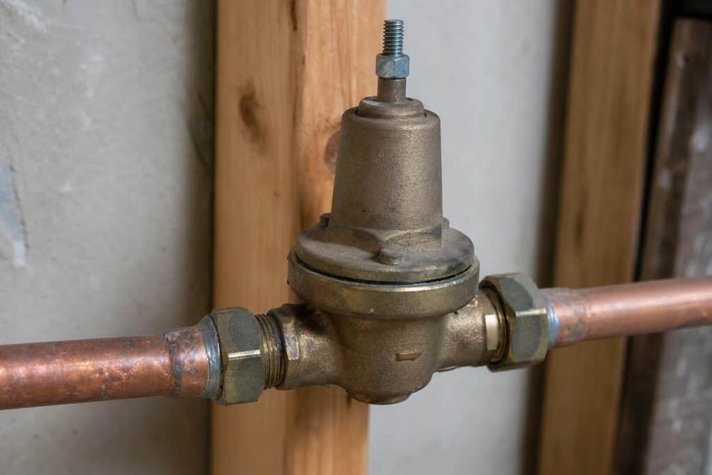 Brass water pressure regulator installed between two copper pipes mounted on wooden supports against a wall.