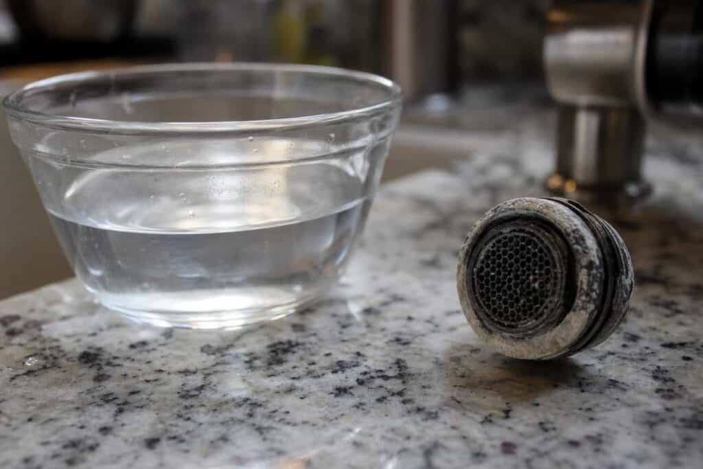 A glass bowl of clear water sits on a granite countertop next to a faucet aerator covered in white mineral deposits.