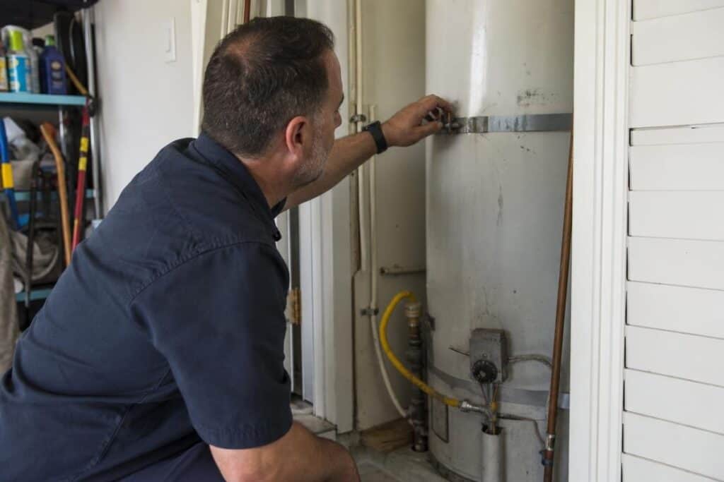 A man in work attire inspects or adjusts a water heater in a utility closet.