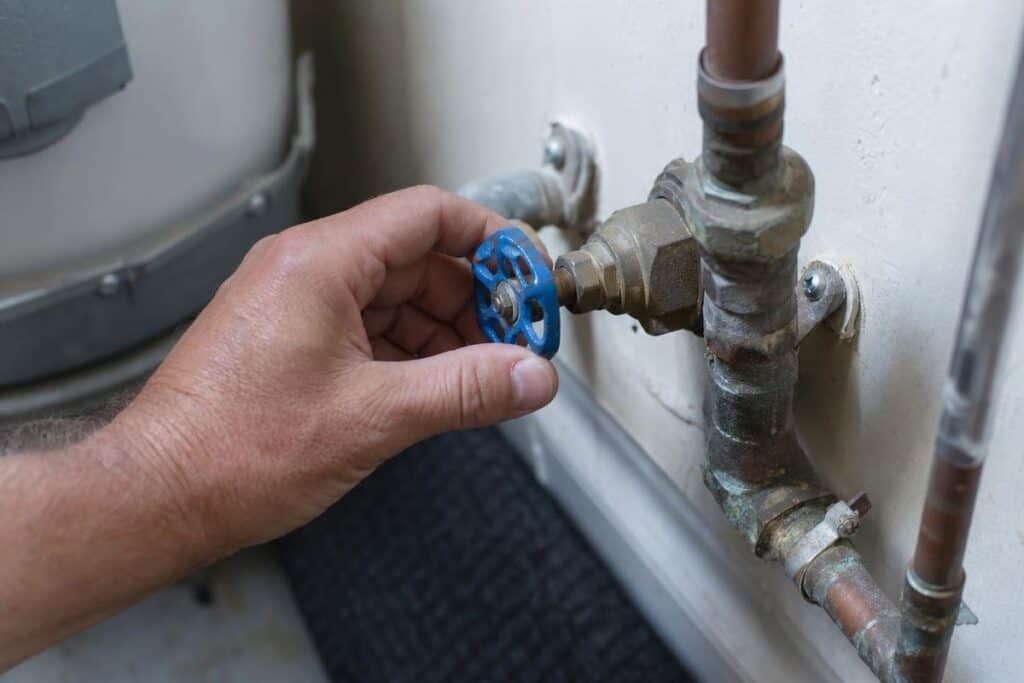 A hand turns a blue metal valve on a set of connected water pipes attached to a wall.