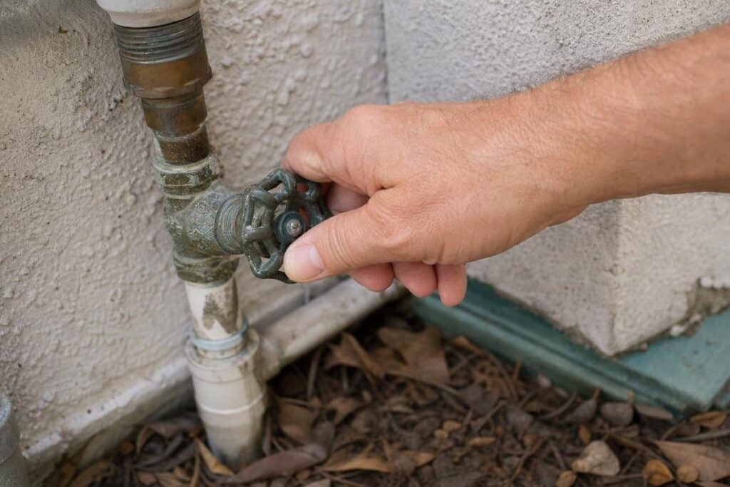 A hand turns an outdoor metal water valve attached to pipes next to a building wall, with dried leaves on the ground below.