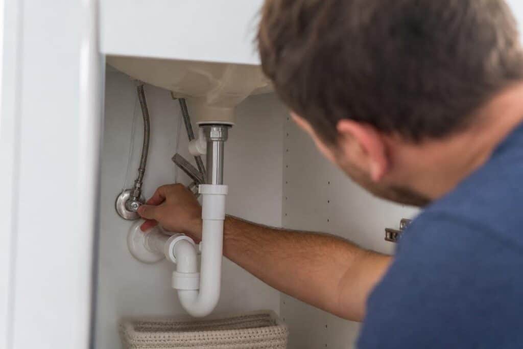A person is tightening a pipe connection under a sink, working on plumbing with a wrench inside a cabinet.