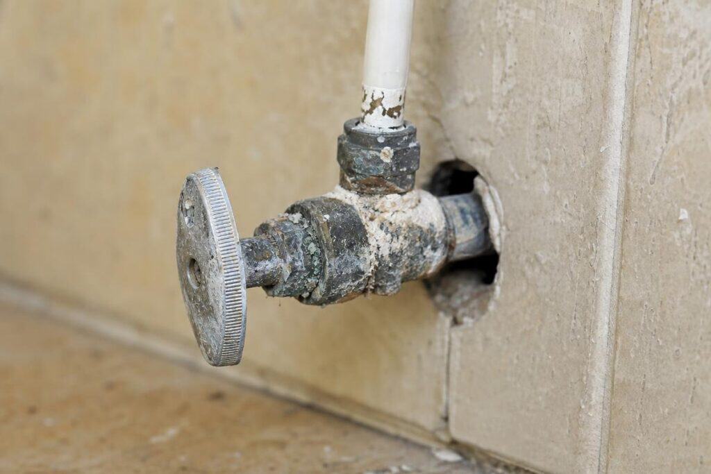 Close-up of a corroded metal water shut-off valve connected to a pipe emerging from a hole in a tiled wall.
