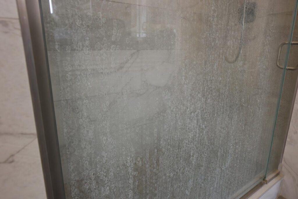 A glass shower door covered in water spots and soap scum buildup, with a metal frame and tiled walls visible in the background.