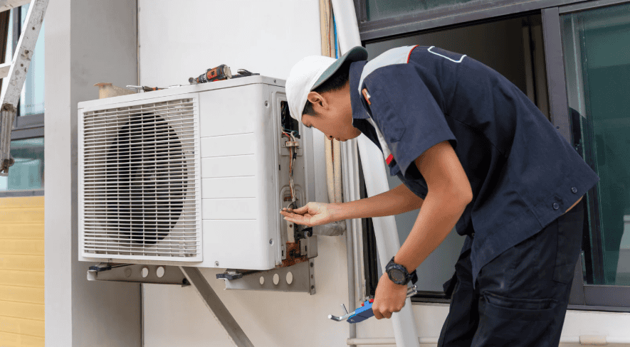 A technician in uniform repairs an outdoor air conditioning unit attached to a building wall, holding a screwdriver and a wrench.