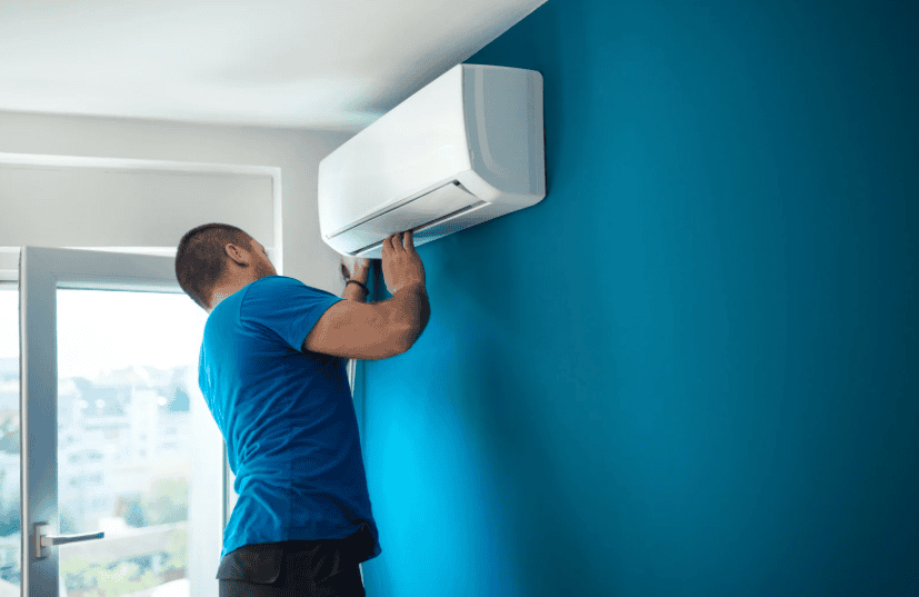 Person wearing a blue shirt installs or repairs a wall-mounted air conditioning unit in a room with blue walls and a window.