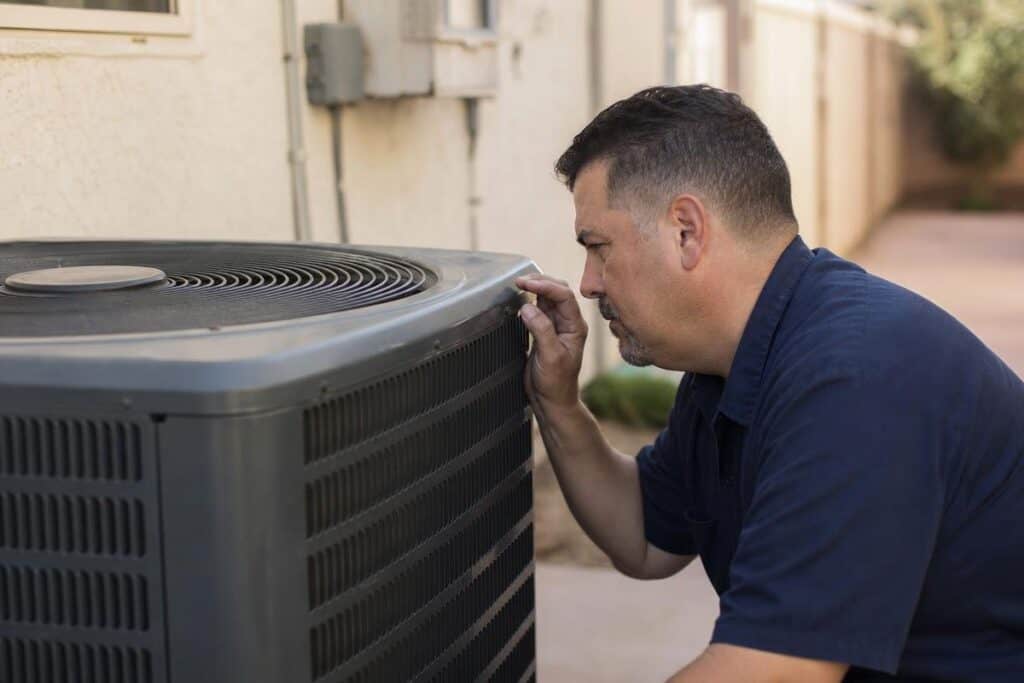A technician inspects an outdoor air conditioning unit next to a residential building during daylight.