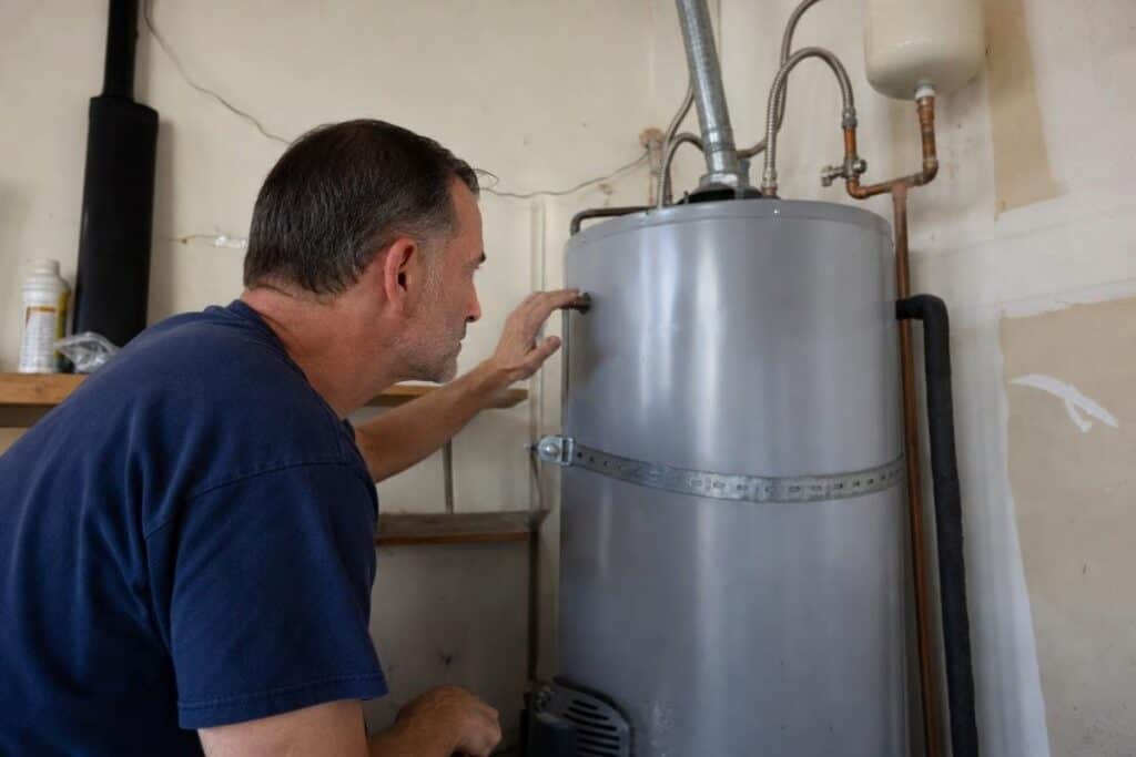 A man in a navy shirt inspects a water heater in a garage, touching the side of the tank and looking at its connections.