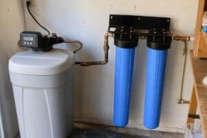 Water filtration and softener system installed on a wall, featuring two large blue filter housings and a control unit with connected pipes.