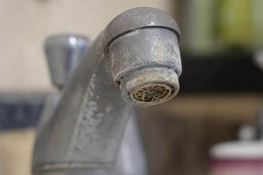 Close-up of a metal faucet with visible mineral buildup and limescale deposits around the spout, indicating hard water residue.