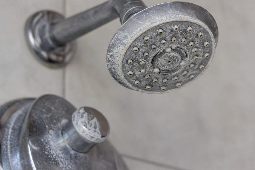 Close-up of a metal showerhead and faucet handle with visible white mineral deposits and limescale buildup.