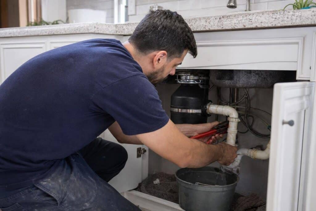 A man in a navy shirt uses a pipe wrench to fix plumbing under a kitchen sink, with a bucket placed below the pipes.