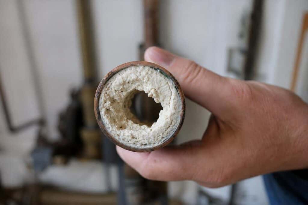 A hand holds a pipe with heavy white mineral buildup inside, restricting the flow through the center of the pipe.
