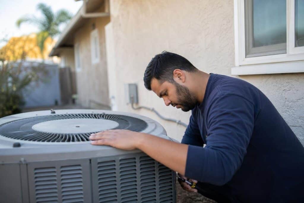 A person inspects or repairs an outdoor air conditioning unit next to a house during the daytime.